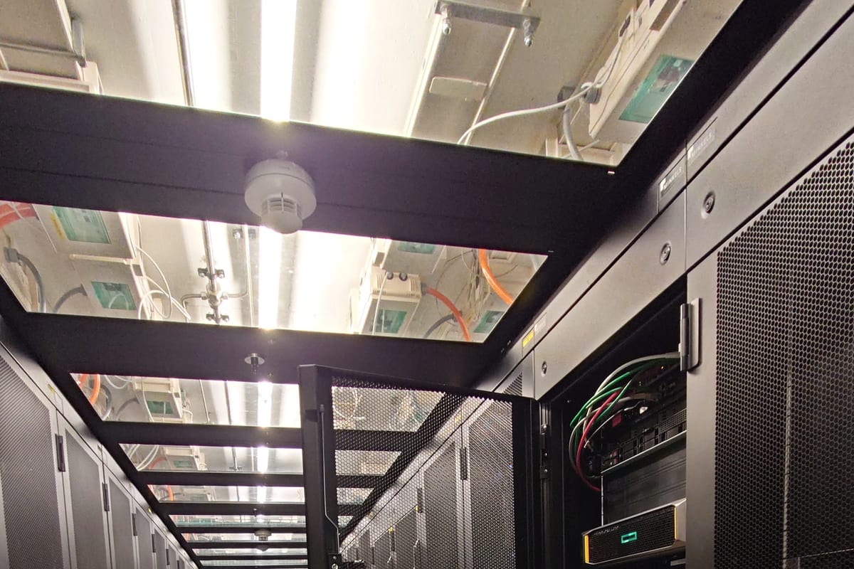 Allée froide confinée dans une salle serveurs : deux rangées de baies noires à portes perforées, verrières de confinement et chemins de câbles au plafond, et une baie ouverte laissant voir les serveurs et leur câblage.